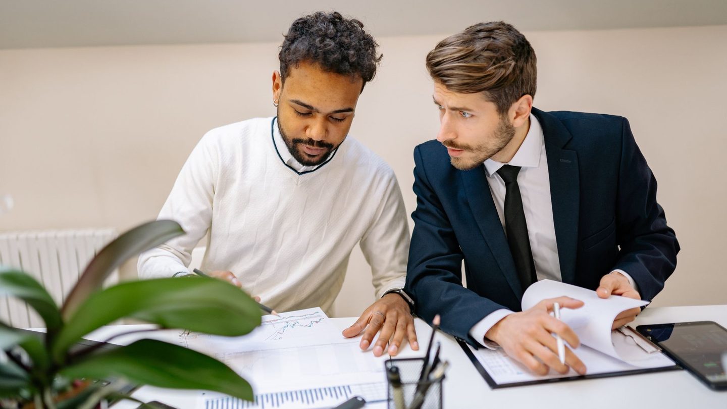 Two colleagues at a desk reading printed schedules, one with a pen, the other turning the pages of a file